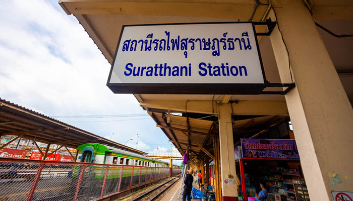 Sign for Suratthani Station at Phunphin Railway Station with a train in the background.